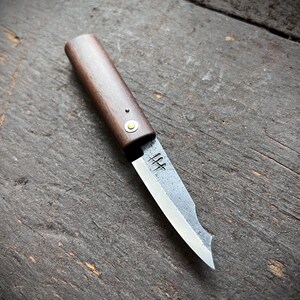 May include: A small fixed-blade knife with a dark brown wooden handle and a single brass rivet. The blade features a textured, dark gray finish with a sharp, polished edge and a distinct maker's mark stamped near the bolster area of the blade.