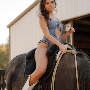 May include: A denim-look bodysuit with a keyhole neckline and ruffle trim. The model is wearing white cowboy boots and riding a gray horse with a black saddle. The background includes a white building and a wooden fence.