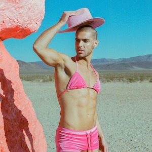 May include: A person wearing a pink velvet bikini top and matching shorts. They are standing in a desert landscape with a large pink rock behind them. They are holding a pink hat in their hand.