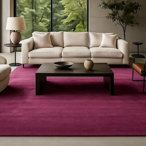 May include: A modern living room with a magenta rug, a beige sofa, and a brown leather chair. A dark wood coffee table holds a bowl and a vase. A round beige chair and a table lamp are also visible.