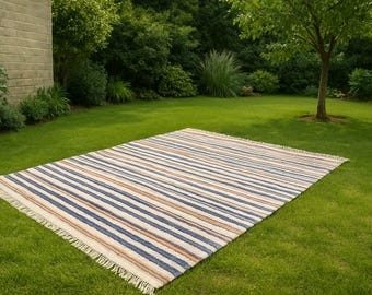 Könnte beinhalten: Rechteckiger Outdoor-Teppich mit Streifen in Blau, Beige und Weiß. Der Teppich liegt auf einem grünen Rasen und hat einen Fransenrand. Im Hintergrund befinden sich üppiges Grün und eine Backsteinmauer.