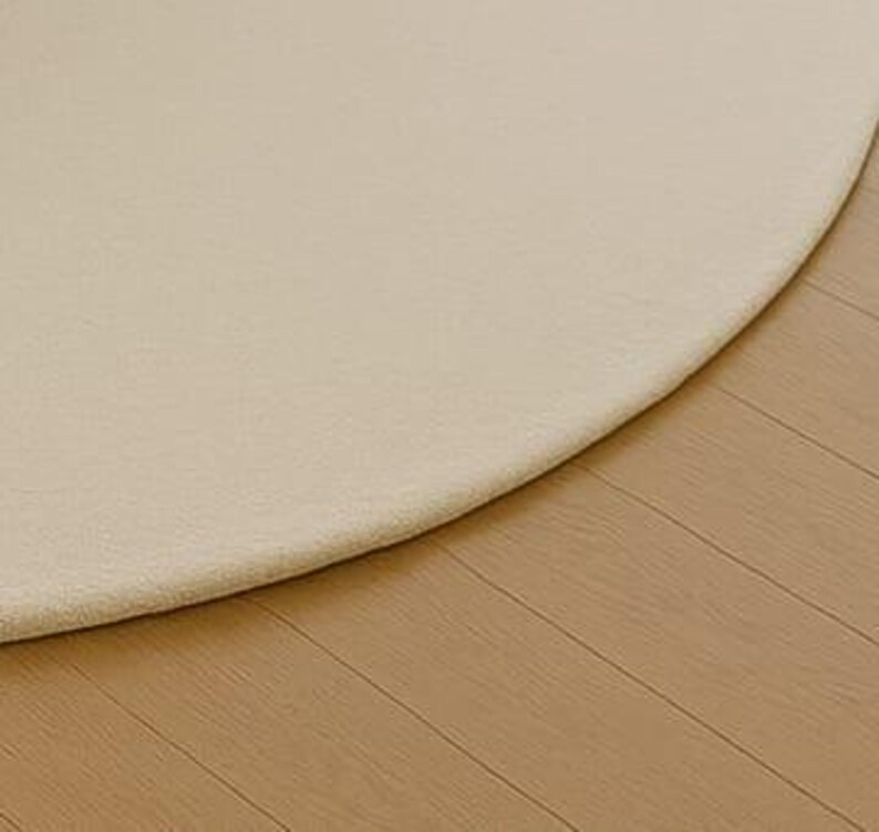 A modern white round rug placed in a beige living room beside large windows with pool and garden view. The soft wool-blend carpet sits under a marble coffee table and cream sofa, creating a bright, elegant, and minimalist interior. Perfect for contemporary, Scandinavian, or neutral-style homes.