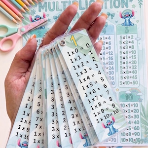 Pode incluir: Um conjunto de cartões de multiplicação plastificados num anel de metal, com o texto 1x0=0 a 1x10=10, decorados com ilustrações de personagens azuis. Os cartões são exibidos sobre uma tabela de multiplicação com lápis de cor.