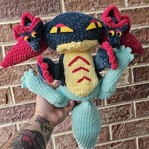 May include: A handmade crochet plush toy in the shape of a creature. The toy features a dark blue head, yellow belly with red markings, and light blue arms and tail. It has red and dark blue accents and yellow eyes.