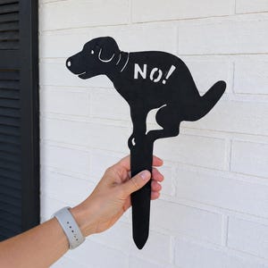 May include: A black metal garden stake shaped like a dog in a squatting position with the word NO! cut out from its side. The silhouette features a pointed bottom end for insertion into the ground and a detailed dog head with a visible eye and ear.