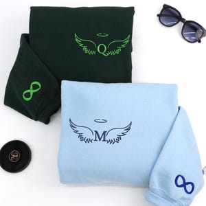Forest green and light blue sweatshirts with angel wing embroidery at the center. The green one features neon green thread with an infinity sign on the sleeve, while the blue one is stitched in navy thread with a matching infinity symbol — a perfect pair for a symbolic duo.