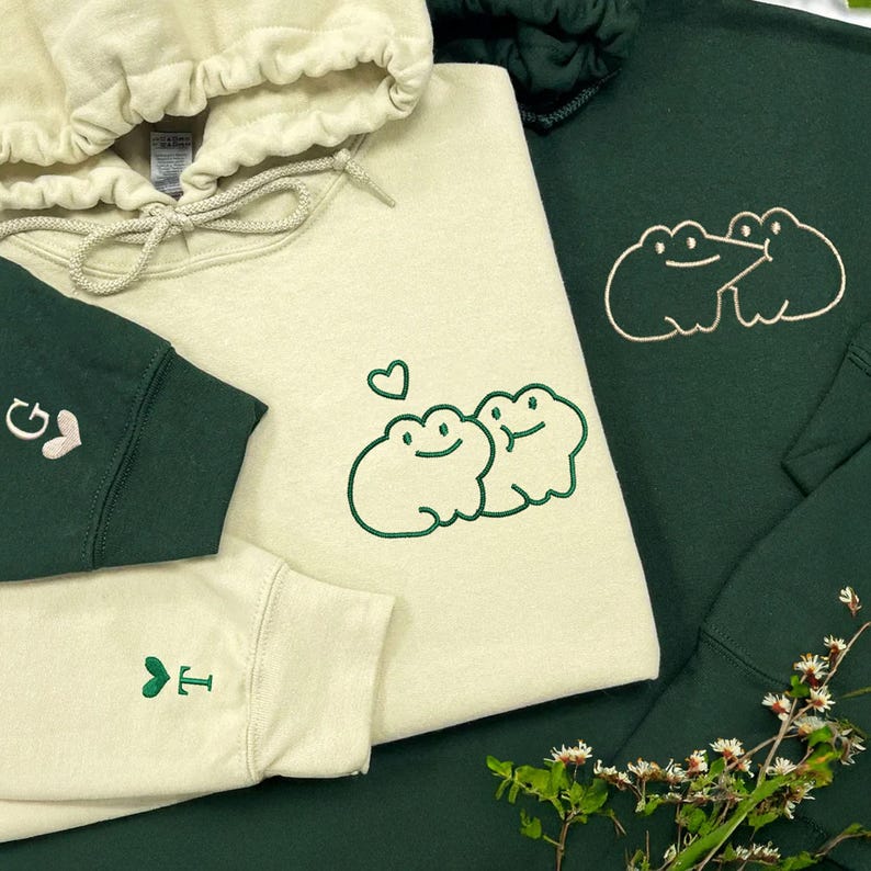 Sand hoodie embroidered on the middle chest with two cute frogs cuddling under a small heart, stitched in green thread. Forest green hoodie embroidered on the middle chest with two frogs facing each other, stitched in light beige thread. Both sleeves include small matching embroidery details with initials and hearts.
