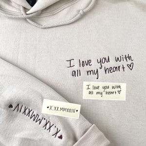 Puede incluir: Una sudadera con capucha beige con cordón y mangas largas. El texto "I love you with all my heart" está escrito en la parte delantera. La manga tiene el texto "X.XX.MMXXIV" y un símbolo de corazón.