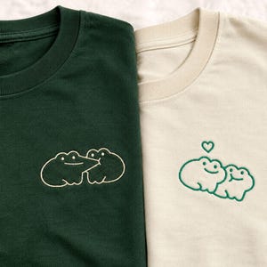 May include: A dark forest green t-shirt and a cream-colored t-shirt, each featuring a simple line-art embroidery of two frogs. The green shirt shows the frogs touching noses, while the cream shirt shows the frogs with a small green heart above.