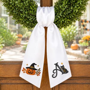 May include: A white satin ribbon hanging from a green wreath. The left tail features an embroidered orange pumpkin wearing a black witch hat with autumn leaves. The right tail displays a large black letter A with a small black cat sitting beside it.