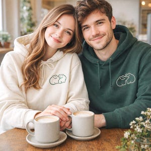 Matching sand and forest green hoodies styled on a couple, featuring a small frog couple embroidery in forest green thread. The simple linework keeps the design cute and minimal. The embroidery placement is subtle, perfect for everyday wear or a cozy coffee date.