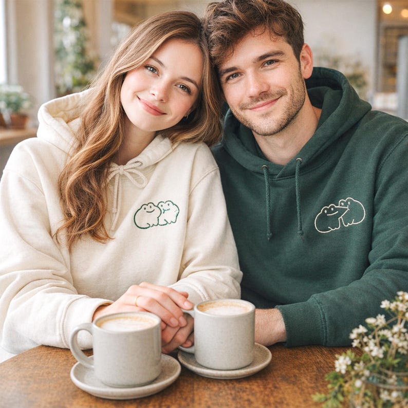 Matching sand and forest green hoodies styled on a couple, featuring a small frog couple embroidery in forest green thread. The simple linework keeps the design cute and minimal. The embroidery placement is subtle, perfect for everyday wear or a cozy coffee date.