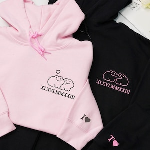 May include: Two hoodies, one pink and one black, are displayed. The pink hoodie features a frog design with the roman numerals XI.XVI.MMXXIII and a heart. The black hoodie has a similar frog design and roman numerals.