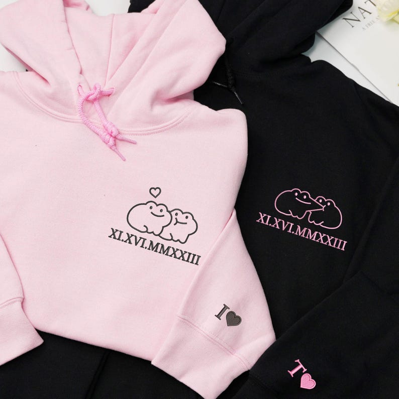 May include: Two hoodies, one pink and one black, are displayed. The pink hoodie features a frog design with the roman numerals XI.XVI.MMXXIII and a heart. The black hoodie has a similar frog design and roman numerals.