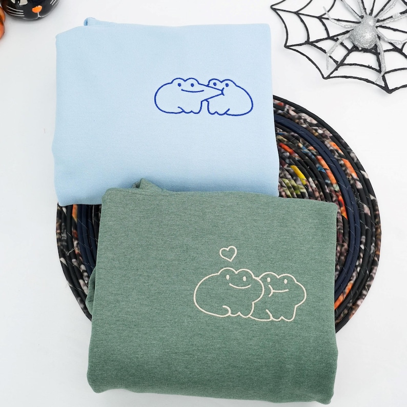 May include: Two sweatshirts, one light blue and one green, are displayed. The light blue sweatshirt features an embroidered outline of two smiling frog-like creatures. The green sweatshirt has a similar design, with a heart above the frogs. The sweatshirts are folded.