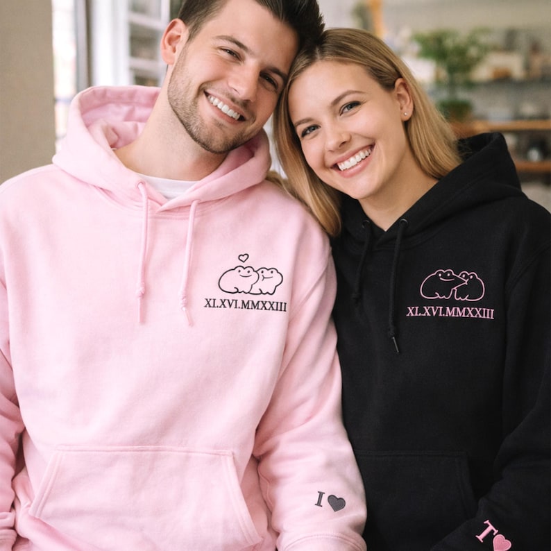 A light pink hoodie and a classic black hoodie showcase the same adorable frog couple embroidery on the left chest. The frogs are outlined in black and soft pink thread, creating a playful contrast. A Roman numeral date beneath the design adds a meaningful, custom detail.
