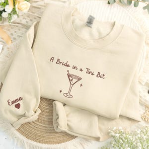 May include: Cream-colored sweatshirt with the text "A Bride in a Tini Bit" and a martini glass graphic. The left sleeve has the name "Emma" and a heart. The sweatshirt is folded on a woven mat with floral accents.