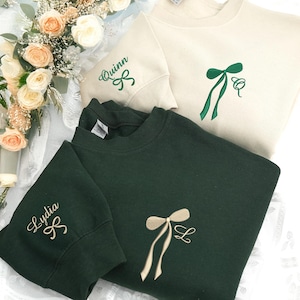 Può includere: Sono mostrate tre felpe, due verdi e una color crema. Ognuna ha un fiocco ricamato e un nome o un'iniziale. La felpa color crema ha un fiocco verde e il nome "Quinn". Le felpe verdi hanno un fiocco beige e il nome "Lydia" e l'iniziale "Q".