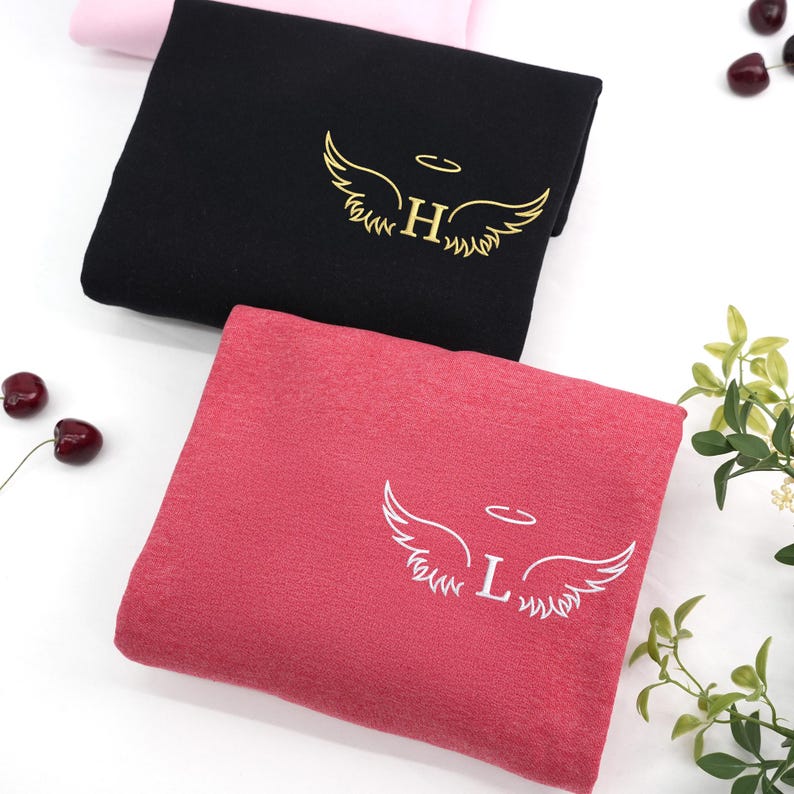 Black and heather scarlet red sweatshirts embroidered with angel wings and halo designs. The black one features gold thread with the letter “H,” while the red one showcases a white “L” — elegant and bold combinations that stand out beautifully.