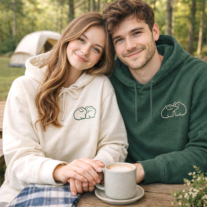 Matching sand and forest green hoodies styled on a couple, featuring a small frog couple embroidery in forest green thread. The simple linework keeps the design cute and minimal. The embroidery placement is subtle, perfect for everyday wear or a cozy coffee date.