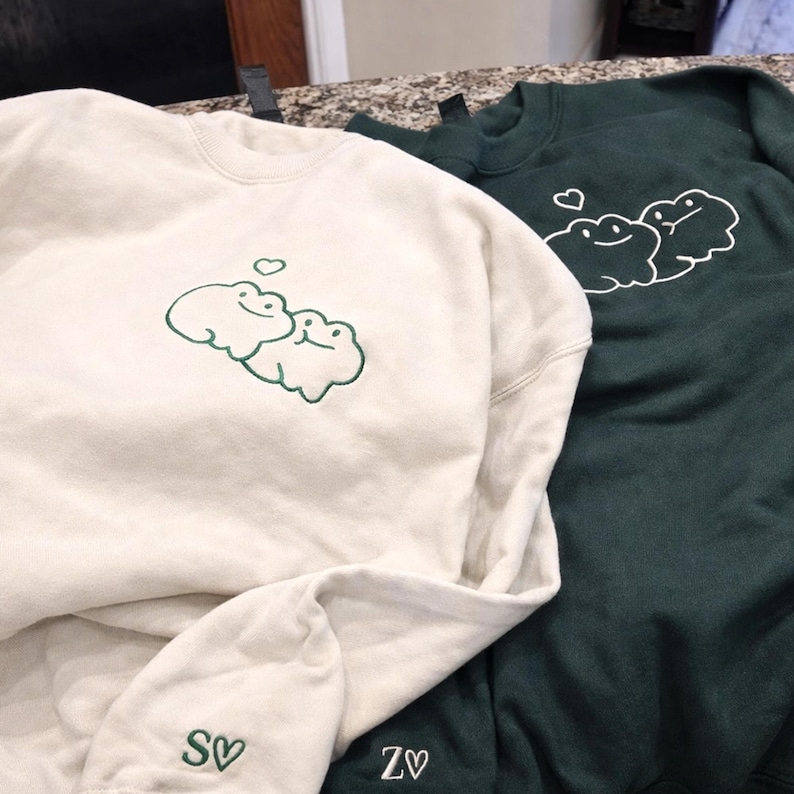 A soft sand sweatshirt paired with a forest green sweatshirt, both featuring a minimalist frog couple embroidery. The design is stitched in clean green and white thread, with a tiny heart detail above the frogs. Subtle sleeve initials add a personalized finishing touch.