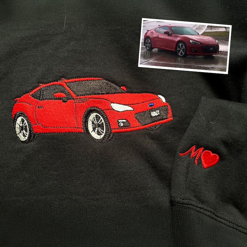 A black sweatshirt featuring a detailed red embroidered car on the chest, replicating the red sports car shown in the attached photo. On the sleeve, there's a custom red embroidery of the letter “M” followed by a heart symbol.