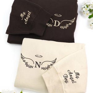 dark chocolate and sand sweatshirts embroidered in cream and dark brown threads. The “D” and “N” initials are paired with phrases like “My Angel” and “My Angel Boy” on the sleeves, creating a heartfelt, cozy tribute design.