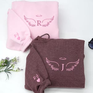 Soft light pink and heather dark maroon hoodies featuring delicate pink embroidery. Each piece has angel wings with initials “R” and “J” on the chest, complemented by matching heart-and-initial details on the sleeves — a romantic, personalized touch.