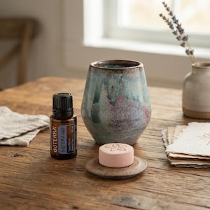 Puede incluir: Un frasco de aceite esencial dōTERRA de vidrio marrón con tapa negra, un recipiente de cerámica moteado en azul verdoso y rosa, y un pequeño disco redondo rosa con una impresión de flor sobre una tapa de cerámica a juego.