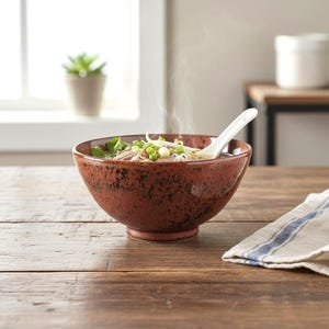 May include: A rustic, speckled brown ceramic bowl filled with steaming soup, garnished with green onions and sprouts. A white spoon rests in the soup. The bowl sits on a wooden table, with a striped cloth napkin in the foreground.