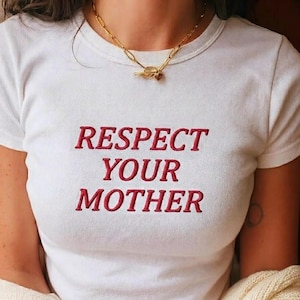 May include: White t-shirt with the embroidered text "RESPECT YOUR MOTHER" in red. A gold-colored chain necklace with a toggle clasp is worn around the neck. The shirt is short-sleeved.