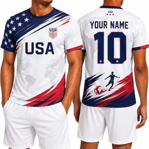 May include: White soccer jersey with blue and red accents, featuring the word "USA" and a shield logo. The jersey has a map design and stars on the sleeves. The back displays "YOUR NAME" above the number "10" and a soccer player graphic.