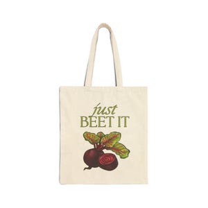 May include: Beige tote bag with the phrase "just BEET IT" in green text. The bag features a watercolor illustration of a beet with green and red leaves. The tote bag has a simple design with long handles.