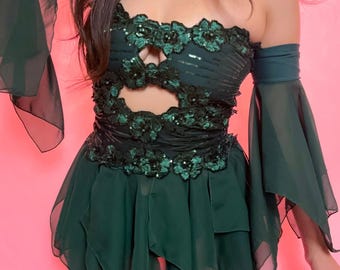 May include: A dark green strapless dress with floral embellishments and a cutout design. The dress features a layered skirt and sheer sleeves. The background is a solid pink.