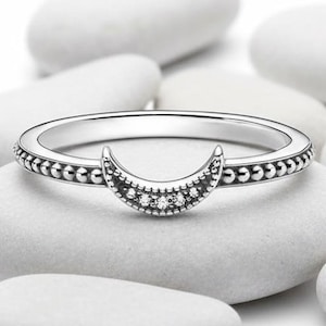 Puede incluir: Anillo plateado con diseño de media luna adornado con pequeñas piedras transparentes. El anillo tiene una banda con cuentas y se presenta sobre piedras blancas lisas, evocando un tema celestial.
