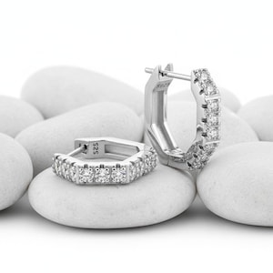 May include: A pair of silver hoop earrings. Each earring has a geometric shape, set with small, clear gemstones. The earrings are displayed on white, smooth stones, creating a clean and elegant presentation.