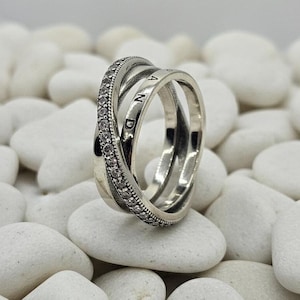 May include: A silver ring with three intertwined bands. One band is encrusted with small, clear stones. The word "AND" is engraved on one of the bands. The ring is set against a background of white stones.
