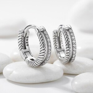 May include: A pair of silver hoop earrings featuring a textured rope-like design on one side and a row of small clear stones on the other. The inner band of the earring is clearly stamped with the text S925, indicating the silver composition.