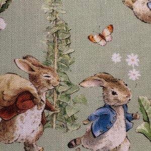 May include: A light green fabric featuring illustrations of rabbits, butterflies, and flowers. One rabbit wears a blue jacket. The illustrations are in a storybook style, with a whimsical and pastoral theme.