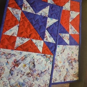 May include: Two quilted placemats with a patriotic theme. The top section features red and blue triangles, while the bottom section showcases a white background with eagle and star patterns. The edges are trimmed with a blue border.