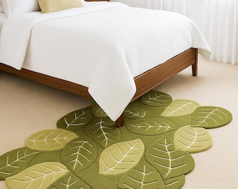 May include: A green leaf-shaped rug with varying shades of green and white leaf vein details. The rug is placed on a beige carpet in a bedroom setting, with a wooden bed frame and white bedding visible in the background.