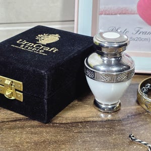 May include: A black velvet box with gold hardware and the text "UrnCraft STUDIO" next to a silver and white urn. A gold bracelet and a strand of black and white beads are also visible.