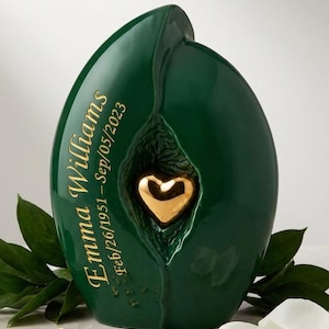 May include: A dark green ceramic urn shaped like a leaf with a glossy finish. It features a central gold-colored heart emblem and the engraved text Emma Williams Feb/26/1951 - Sep/05/2023. The design is smooth with a curved, organic silhouette.
