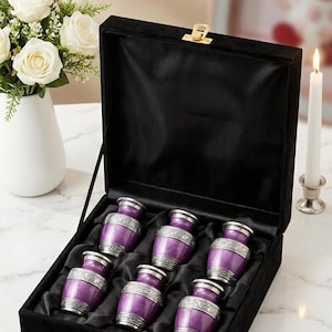 May include: A set of six small, purple urns with silver trim, presented in a black velvet-lined box. The urns are arranged neatly inside the box, which is closed with a gold clasp. A bouquet of white roses and a lit candle are in the background.