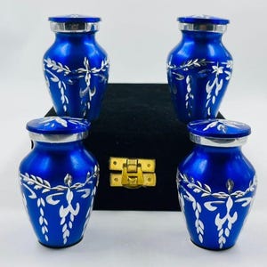 May include: Four cobalt blue keepsake urns with silver lids and floral patterns. The urns are displayed in front of a black box with a gold clasp. These small, decorative urns are likely for storing cremated remains.