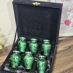 May include: A set of six small, green and black decorative urns with silver-colored lids, presented in a black velvet box. The urns have a floral pattern. The box is closed with a gold-colored clasp.