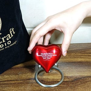 May include: A red heart-shaped memorial with engraved text. The heart is mounted on a silver ring base. The text reads "Erica McClure Moore September 21, 1976 - December 20, 2019" and "Not Gone Just A Little Ahead... Waiting."
