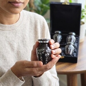 May include: A small, dark brown metal keepsake urn with a silver-toned lid and an engraved white leaf pattern. The urn is cylindrical with a rounded body. In the background, a black velvet-lined display box contains three additional matching urns.