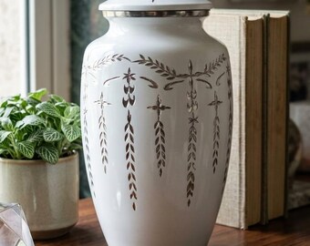 May include: A white ceramic urn with a lid, featuring a decorative pattern of stylized leaves and crosses in a metallic silver color. The urn is displayed on a wooden surface.