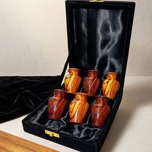 May include: A set of six small, polished, amber-colored urns with swirling wood-grain patterns, arranged in a black velvet-lined display box. The box features a gold-tone metal latch and a black satin interior lining to hold the six individual vessels.
